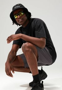 Schwarzer Bucket Hat, reflektierende Sonnenbrille, schwarzes kurzärmeliges Hemd, graue Shorts, schwarze Sneakers und strukturierte Haut, in einer hockenden Position posiert.