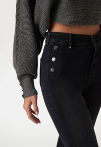 Black high-waisted jeans feature a smooth texture, silver hardware accents, and a fitted design. A gray ribbed sweater is partially visible.
