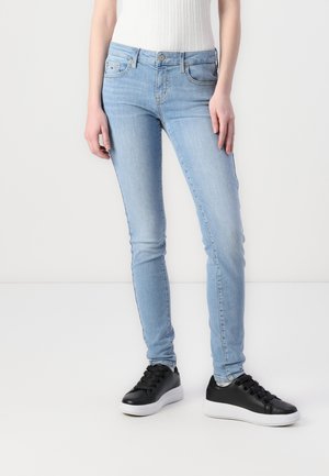 Personne portant un jean skinny bleu clair et des baskets noires à semelles blanches, debout devant un fond blanc uni.
