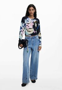 Ilgas rankovėmis raštuotas tamsiai mėlynas viršus su gėlių raštais, derinamas su šviesiai mėlynais, plačiomis kojomis, aukštu liemeniu džinsais. Modelis laiko juodą delninę.