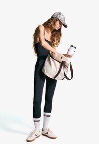 Schwarzer, taillierter Jumpsuit, gemusterte Mütze, Canvas-Tasche mit dunklen Griffen, gestreifte Socken und weiße Sneaker. Hält einen Kaffeebecher und greift in die Tasche.