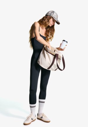 Black fitted jumpsuit, patterned cap, canvas bag with dark handles, striped socks, and white sneakers. Holding a coffee cup while reaching into bag.