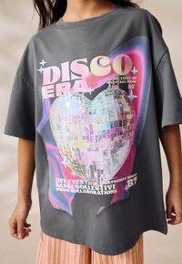 Gray oversized T-shirt featuring a colorful disco heart graphic with sequins, and bold text, paired with striped pastel pants.