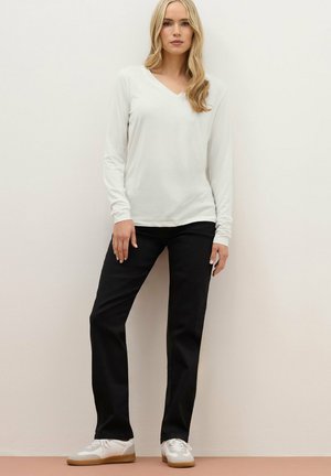 Mujer rubia de pie contra la pared, vistiendo una blusa blanca de manga larga con escote en V, pantalones negros y zapatillas blancas con suela de goma.