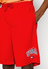 Pantaloni corti sportivi rossi con vita elastica, tasche anteriori e un grande logo "TOMMY USA JEANS" in bianco e blu sul lato inferiore.