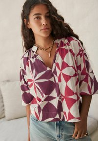 Chemise multicolore à manches courtes présentant un motif géométrique dans des nuances de violet et de rouge sur un fond clair. Conception en V.