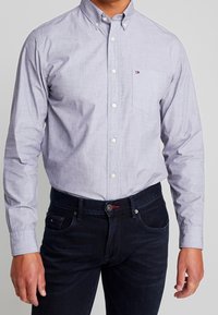 Vaaleansininen nappipaita, jossa on tasku ja teksturoidusta kankaasta valmistettu, yhdistetty tummansinisiin farkkuihin, joissa on punainen yksityiskohta napissa.