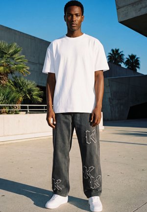 Jeune homme debout en extérieur portant un t-shirt blanc uni, un jean large et foncé avec des motifs patchwork, et des baskets blanches sous un ciel clair.