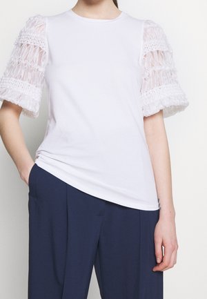 Haut blanc à manches courtes avec manches bouffantes texturées et semi-transparentes, associé à un pantalon plissé bleu foncé, main dans la poche.
