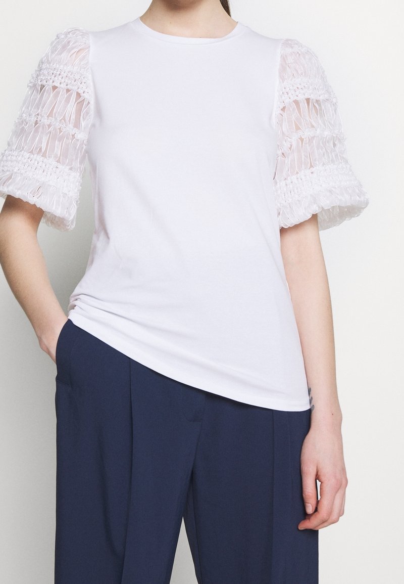 Haut blanc à manches courtes avec manches bouffantes texturées et semi-transparentes, associé à un pantalon plissé bleu foncé, main dans la poche.