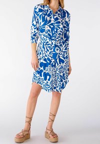 Abito camicia a fiori blu e bianchi con maniche lunghe, vita sottolineata da una cintura e taglio fino al ginocchio. Mostrati un paio di sandali con plateau a fasce beige.