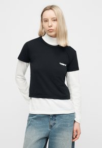 Ξανθιά γυναίκα με τζιν ανοιχτού χρώματος φοράει μαύρο κοντό T-shirt με λευκό λογότυπο "OBEY" πάνω από λευκό ζιβάγκο με μακριά μανίκια, μπροστά σε απλό φόντο.