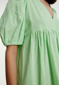 Robe vert clair à manches bouffantes avec un décolleté en V, tissu texturé et jupe froncée, mettant en valeur une silhouette fluide et décontractée.