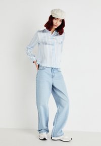 Camisa de botão azul-claro riscada, com mangas compridas, combinada com jeans largos de lavagem clara e ténis brancos com detalhes pretos.