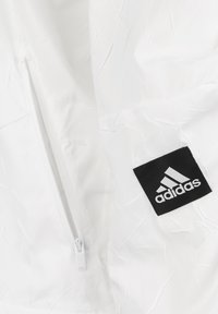 White jacket featuring a textured fabric, a zippered pocket, and a black Adidas logo label on a contrasting background.