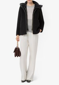 Giacca nera con cappuccio, chiusura a zip e dettagli dorati. Maglione a righe grigio chiaro sotto. Pantaloni bianchi a gamba larga, borsa marrone.