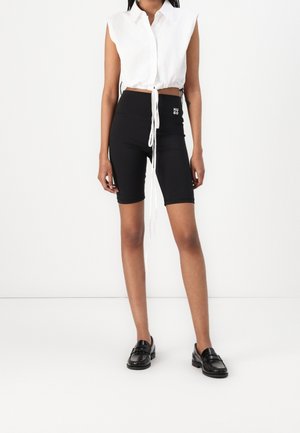 Vrouw draagt een wit mouwloos cropped shirt met strikjes en zwarte high-waisted fietsbroek met "HUGO"-logo, gecombineerd met zwarte loafers.