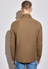 Hnědá, texturovaná košile s dlouhým rukávem, vysokým límcem a rovním lemem, v kombinaci s modrými džíny, viděná zezadu.