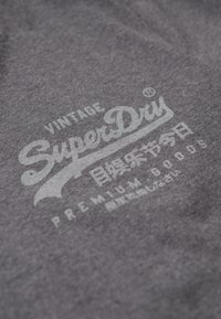 Grå stof med en blød tekstur har hvid trykt tekst "Vintage SuperDry" og yderligere mærker i forskellige skrifttyper og sprog.