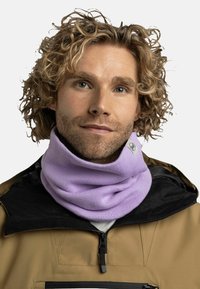 Purple knitted neck gaiter, softly textured, worn over a black jacket with zippered pocket. Features a small logo tag on the side.