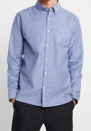 Uomo che indossa una camicia azzurro chiaro con bottoni, maniche lunghe e taschino sul petto, abbinata a pantaloni scuri con fantasia.