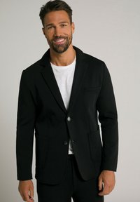 Blazer nero realizzato in morbido tessuto, con revers a punta, due tasche frontali e chiusura a bottone singolo su una t-shirt bianca.