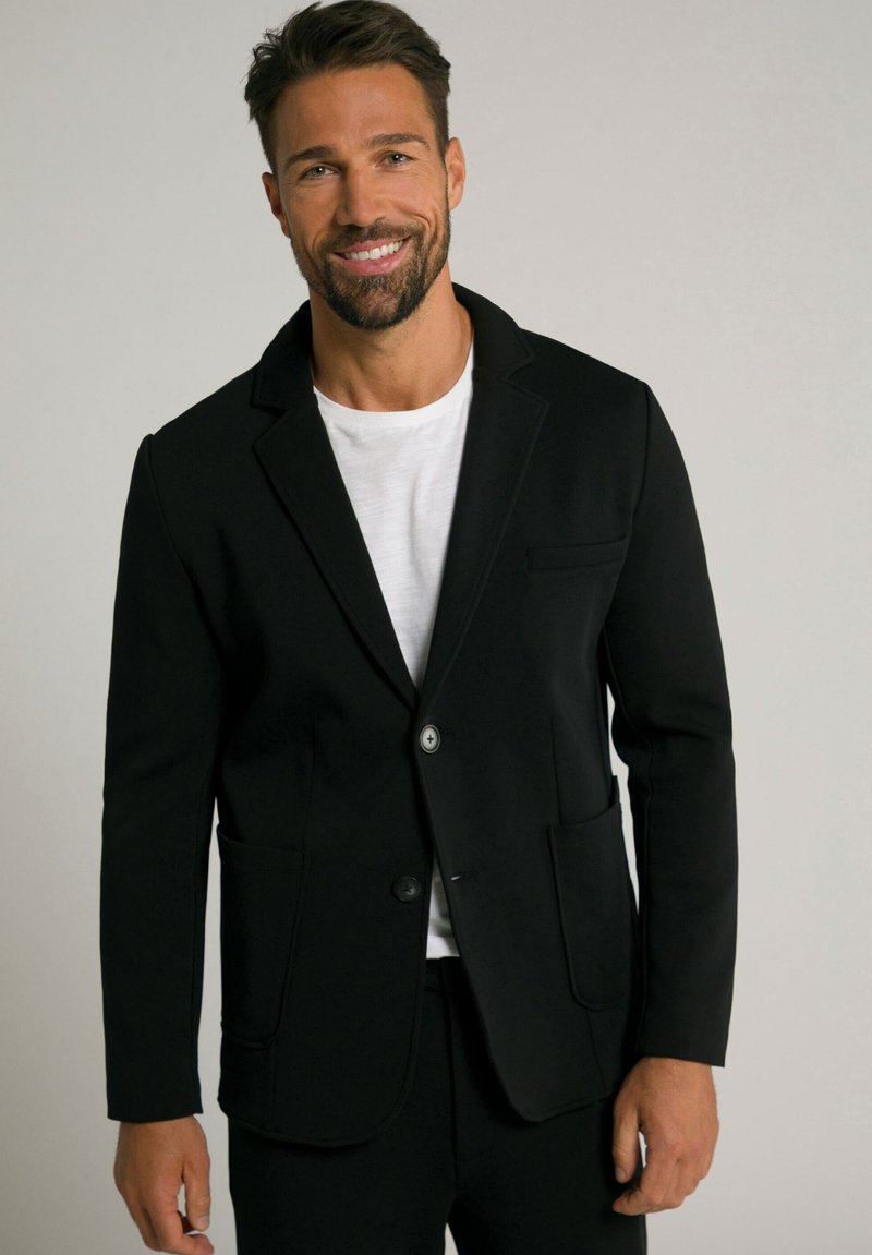 Blazer nero realizzato in morbido tessuto, con revers a punta, due tasche frontali e chiusura a bottone singolo su una t-shirt bianca.