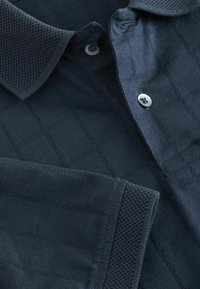 Mørkeblå polo t-shirt med et struktureret diamantmønster og en solid, ribbet krave. Indeholder en knaplukning og syede detaljer.