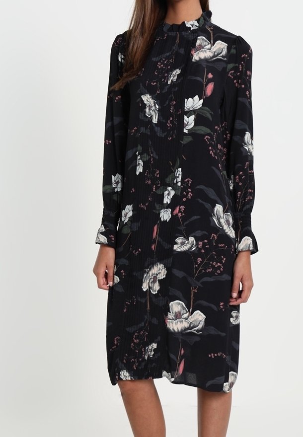 Robe noire à manches longues, longueur genou, avec imprimé floral blanc et rose et col montant à volants portée par une personne aux longs cheveux bruns.