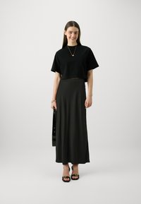 Top negru scurt, fustă neagră lungă și vaporasă până la gleznă și sandale cu toc negre. Bijuterii simple din aur. Textură de material fin, design minimalist.