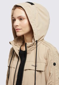 Beige Kapuzenjacke mit schwarzen Kordeln, mit einem Reißverschluss und zwei Brusttaschen. Glattes Material mit einem dezenten Glanz.