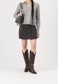 Femme portant un haut gris à manches longues, une mini-jupe en denim noire, des bottes de cow-boy brodées marron, et tenant un sac à bandoulière vert foncé.