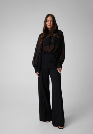 Femme aux longs cheveux portant un chemisier noir transparent à manches bouffantes et un pantalon noir taille haute à jambes larges sur un fond uni.