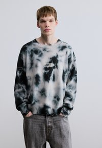 Μπλούζα τύπου tie-dye σε ασπρόμαυρο, με ριμπ γιακά και μανσέτες, και μικρό πατς με το λογότυπο "HUGO" στο μπροστινό μέρος. Φοριέται με γκρι τζιν.