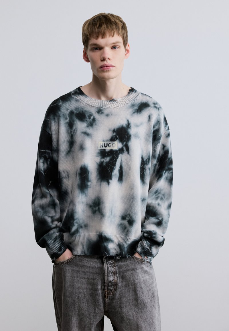 Μπλούζα τύπου tie-dye σε ασπρόμαυρο, με ριμπ γιακά και μανσέτες, και μικρό πατς με το λογότυπο "HUGO" στο μπροστινό μέρος. Φοριέται με γκρι τζιν.