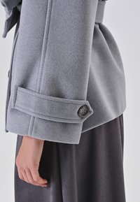 Cappotto di lana grigio chiaro con tessuto texturizzato, dotato di un polsino con bottone e di un design aderente che si allarga leggermente in vita.