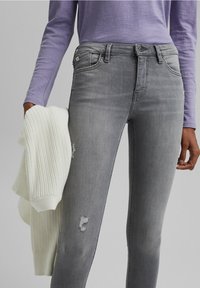 Vaqueros pitillo grises con detalles desgastados, combinados con un suéter blanco de punto acanalado, sostenido en la mano izquierda, y una camiseta de manga larga color lavanda.