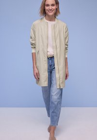 Kvinde smilende, iført beige lang jakke, pink top, lyseblå jeans og beige sko, gående mod en lyseblå baggrund.