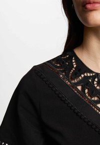 Zwarte jurk met een intrigerende kanten halslijn met uitsnijdingen en golvende afwerking. Gladde stof met delicate details langs de randen.