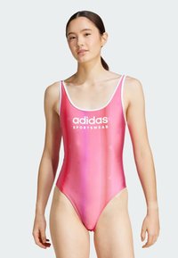 Růžový jednodílný plavky s síťovaným vzorem, obsahující bílé logo "adidas SPORTSWEAR." Tenké ramínka a vysoký střih nohou.