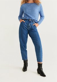 Jasnoniebieski sweter prążkowany, niebieskie dżinsy z wysokim stanem oraz czarne botki za kostkę. Dżinsy mają luźny krój z podwiniętymi końcami.