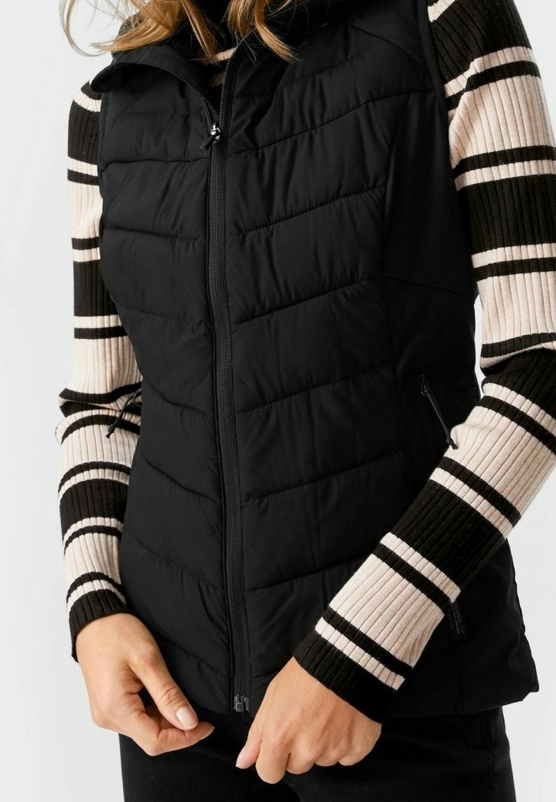 Gilet matelassé noir avec fermeture éclair. Présente des sections matelassées et des poches latérales. Porté par-dessus un haut à manches longues rayé et côtelé en crème et noir.