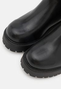 Primer plano de botas de cuero negro con punteras redondeadas y suelas gruesas de caucho texturizado sobre una superficie blanca.