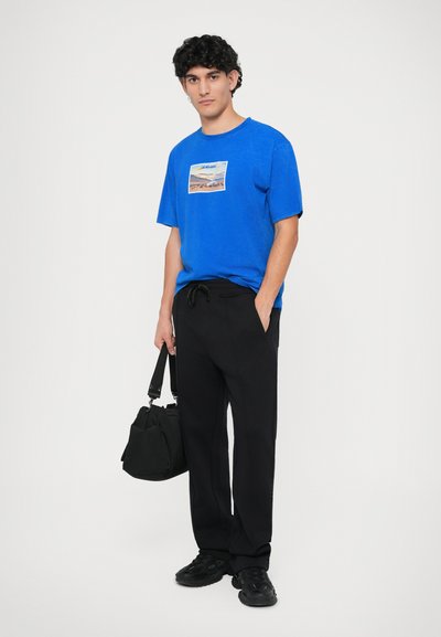 Blue cotton t-shirt with a front graphic and relaxed fit, paired with black drawstring pants and black sneakers, holding a black bag.
