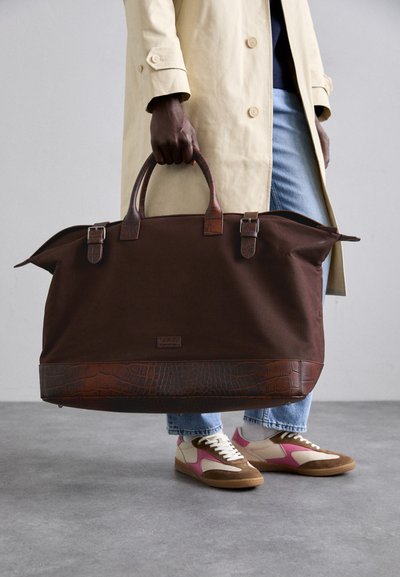 Large brown canvas duffel bag with crocodile-embossed trim, double handles, and silver hardware; worn with light denim and brown sneakers.