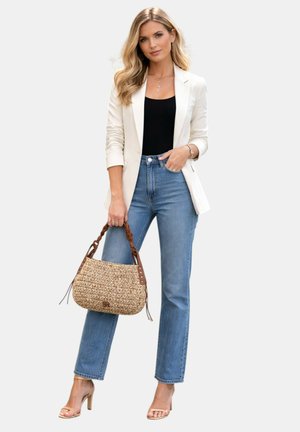 Donna con capelli biondi che indossa un blazer bianco, top nero, jeans blu, tacchi beige, che tiene una borsa intrecciata con manico marrone intrecciato.
