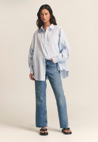 Camicia blu chiaro oversize con bottoni e tasca sul petto, abbinata a jeans blu chiaro a vita alta e sandali neri con la zeppa.