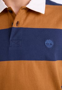 Brah in mornasti polo majica z belo ovratnikom, ki ima gumbasto zapiranje in izšito logo na prsih. Mehka bombažna tekstura.