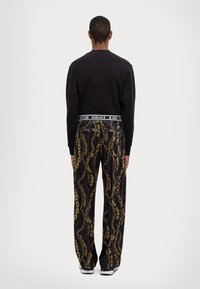 Black sweatshirt with long sleeves, paired with black pants featuring a gold chain pattern and branding. White footwear completes the look.