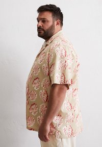 Korte mouwen overhemd in beige stof met grote roze en koraal monstera blad prints. Relaxte pasvorm met een kraag en zijsplit.